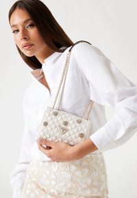 Borsa bianca trapuntata con catena argentata, caratterizzata da decorazioni floreali e un logo triangolare. Appoggiata su una gonna chiara e strutturata.