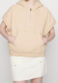 Béžová mikina na zip s velkou přední kapsou, krátkými rukávy a šňůrkami. Obsahuje malé logo "obey" na hrudi.