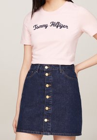 Camiseta de algodón rosa con "Tommy Hilfiger" bordado en azul marino. Combinada con una falda de mezclilla oscura con botones dorados y dos bolsillos.
