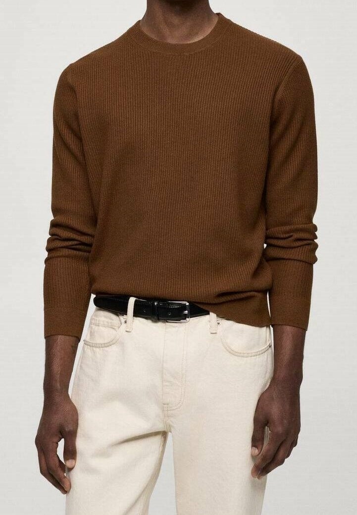 Uomo che indossa un maglione marrone a coste con maniche lunghe infilato in pantaloni bianchi sporco con una cintura nera, in piedi di fronte a uno sfondo neutro.