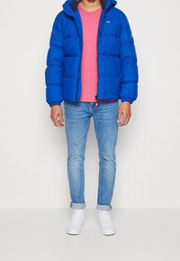 Blue puffer jacket with a hood, featuring horizontal quilted panels and zipper closure. Worn over a pink shirt, paired with light blue jeans and white sneakers.