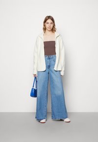 Hvit fleece zip-up jakke, brun tube topp, vide blå jeans, hvite sko og en liten blå veske. Enkelt design med minimale aksenter.
