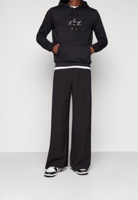 Sort hættetrøje med en frontlomme og snore, med hvid logo tekst. Kombineret med løstsiddende sorte bukser og sorte sneakers med hvide detaljer.