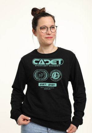 Kobieta w okularach i z koczkem, ubrana w czarną bluzę z turkusowymi logo i napisem "CADET Starfleet Academy", trzymająca dół bluzy i patrząca w prawo.