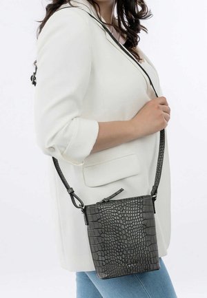 Bolso bandolera negro con textura de cocodrilo, forma plana, correa ajustable y diseño minimalista, sostenido por una persona que lleva un blazer blanco.