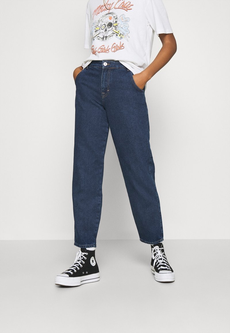 Tmavě modré džínové kalhoty s vysokým pasem, s bočními kapsami a rovným střihem nohavic, doplněné černými teniskami s vysokým svrškem.