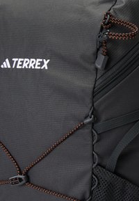 Mochila negra con tejido texturizado. Las características incluyen el logo blanco "TERREX", cordones elásticos acentuados en naranja y detalles de malla.