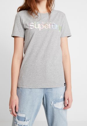 Nuori henkilö pukeutuneena harmaaseen Superdry-paitaan ja vaaleansiniset repaleiset farkut, seisoo käsivarret rentoina sivuilla.