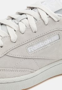 Zapatilla de ante en gris claro con superficie texturizada, cordones gruesos y un parche de logo blanco. Presenta una suela de goma y detalles de costura.