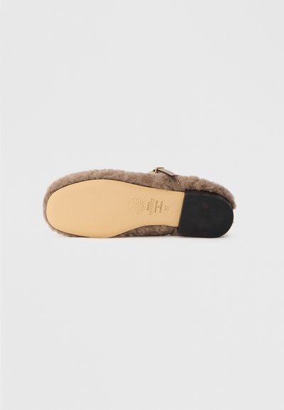 Scarpa slip-on in pelo marrone con punta arrotondata, morbida al tatto e suola piatta. Presenta una piccola cinghia e dettagli in metallo dorato.