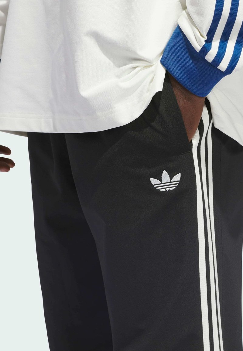 Black athletic pants with white side stripes and a white leaf logo accent, paired with a white shirt featuring blue striped sleeves.