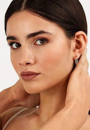 Goldene Creolen schmücken ein Ohr, mit einer glatten Oberfläche und glänzenden Finish. Das Gesicht wird von glattem, dunklem Haar und natürlichem Make-up umrahmt.