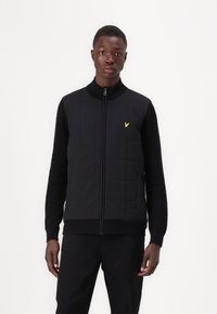 Black quilted vest with a high collar and zipper, featuring textured black sleeves and a small yellow logo on the chest.