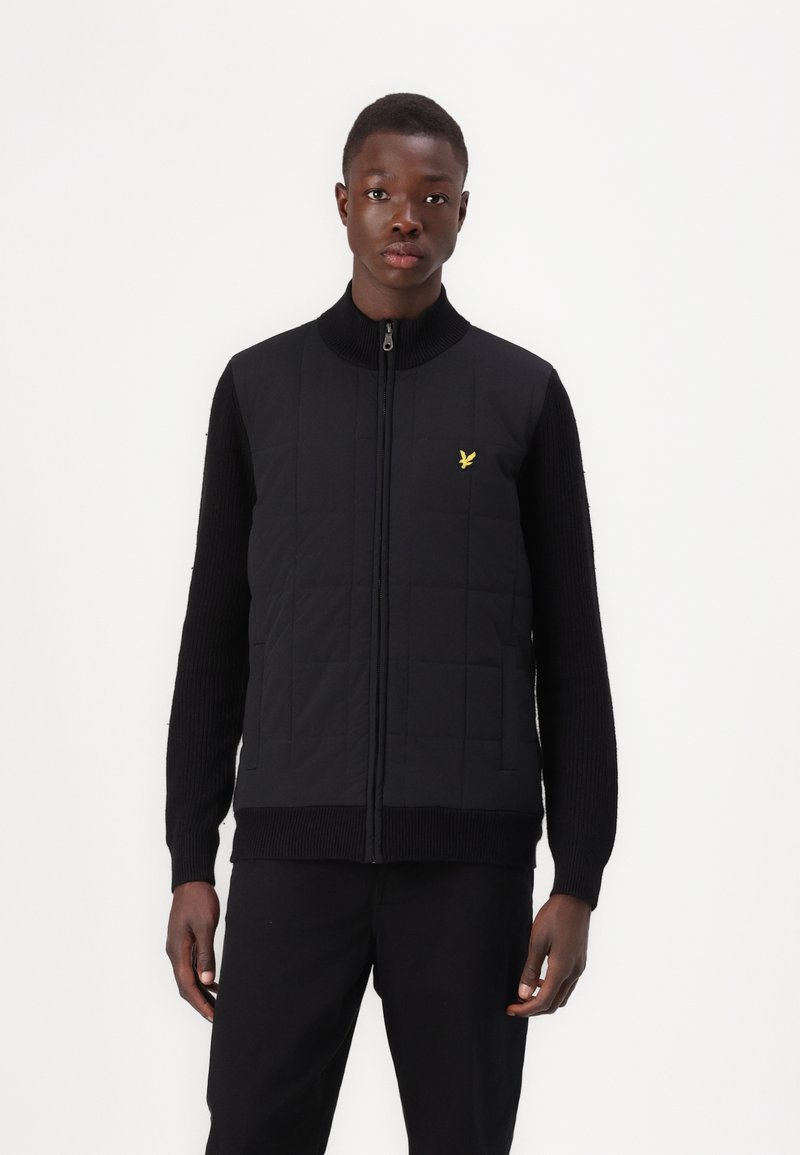 Black quilted vest with a high collar and zipper, featuring textured black sleeves and a small yellow logo on the chest.