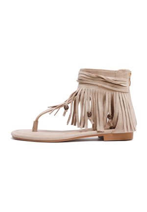 Sandale plate en daim beige avec fermeture éclair à la cheville, détails à franges et perles en bois sur les brides.