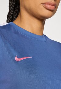 Κόκκινο αθλητικό μπλουζάκι Nike με κοντά μανίκια  και ροζ λογότυπο στο στήθος. Το ύφασμα έχει υφή και εμφάνιση που αναπνέει.