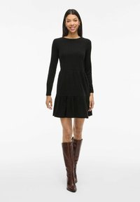 Robe noire côtelée avec des manches longues, jupe à volants et col rond, associée à des bottes en cuir marron jusqu'aux genoux. Fond blanc propre.