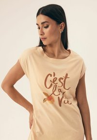 T-shirt w kolorze brzoskwiniowym z krótkim rękawem. Posiada graficzny nadruk z napisem "C'est la Vie" oraz ilustracją brzoskwini. Miękka tekstura.
