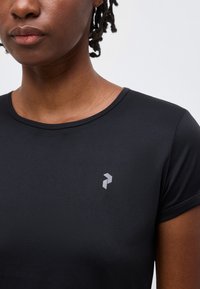 Κοντινό πλάνο ενός ατόμου που φοράει ένα μαύρο αθλητικό κοντομάνικο μπλουζάκι με μικρό γκρι γεωμετρικό λογότυπο στο αριστερό στήθος.