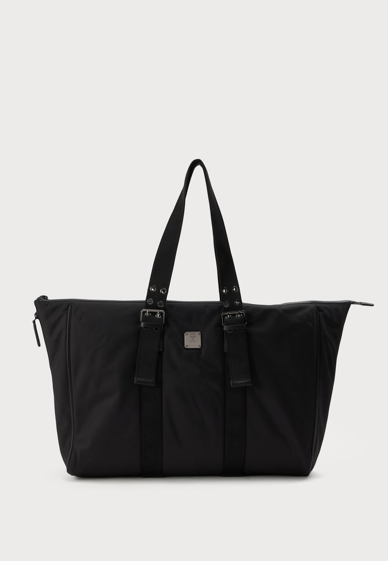 Grand sac cabas noir avec deux poignées, boucles à l'avant et une petite plaque métallique carrée avec logo, présenté sur un fond clair uni.