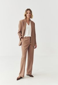 Completo beige su misura con blazer a un bottone e pantaloni a gamba dritta, abbinato a un top bianco a scollo a V e ballerine marroni a punta.