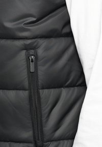 Melns polsterēts bezsuga ar rāvējslēdzēja priekšpusi, kurai ir teksturēta apdare un redzams quiltēts raksts. Rāvējslēdzējs ir melns ar vilkšanas cilpu.