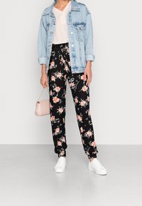 Chaqueta de mezclilla sobre una blusa clara, pantalones negros con flores y puños elásticos, zapatillas blancas y un pequeño bolso rosa claro.