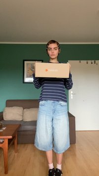 Zalando doboz egy személy által tartva, aki sötétkék csíkos hosszú ujjú felsőt és világoskék farmer rövidnadrágot visel, egy zöld falú szobában.