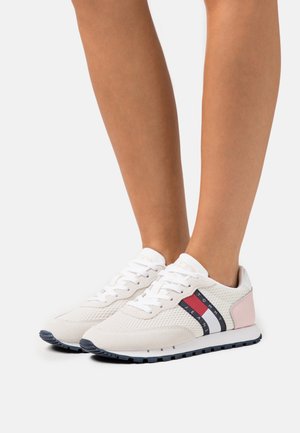 Persona usando zapatillas de malla blanca con acentos rosas en el talón y franjas con el logo de Tommy Jeans en azul marino, rojo y blanco, de pie sobre un fondo blanco.