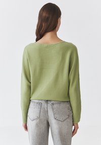Grønne ribbede strikkegenser med en bred udskæring og lange ærmer, der har et asymmetrisk design og en cropped pasform, kombineret med grå jeans.