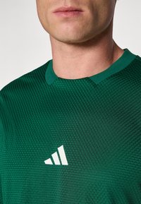 Groene sportshirt van textuurstof; heeft een ronde kraag en een wit logo bestaande uit drie diagonale strepen.