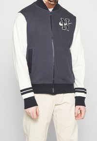 Marineblauwe en witte bomberjack met ritssluiting, geribbelde manchetten, twee zijzakken, voorzien van logoborduring op de borst en gestreepte accenten.