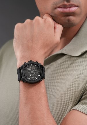 Mand iført et sort robust armbåndsur med flere urskiver på venstre håndled, hånden hvilende på hagen, iført en lyset tekstureret polo.