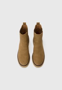 Botas de tobillo de ante en color arena con una textura suave, paneles laterales elásticos, una lengüeta para ayudar a ponerlas y una punta redondeada, que presentan un contorno oscuro en la suela.