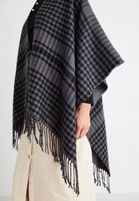 Grau karierter Poncho mit fransigen Kanten, der eine Mischung aus Schwarz und hellgrauen Tönen zeigt, getragen über einem schwarzen Langarmtop und einem beigen Rock.