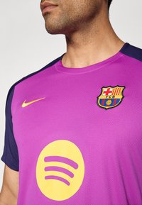 Muž v purpurovom a námorníckom drese FC Barcelona s žltými logami Nike a Spotify, orezaný na brade a hornej časti trupu.