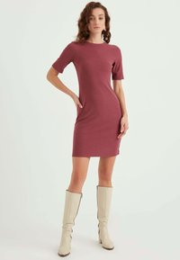 Ribbet, tætsiddende kjole i mauve, med korte ærmer og knælangt snit, parret med høje beige støvler med lynlås på siden.