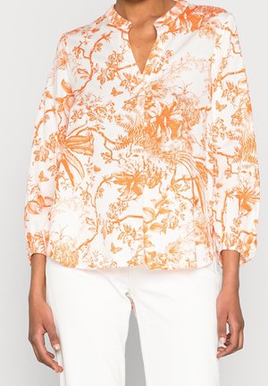 Femme portant un chemisier blanc avec des motifs floraux et de papillons orange et un pantalon blanc, montrée du cou aux hanches avec les bras détendus le long du corps.
