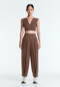 Femme debout, face à nous, portant un haut court plissé marron sans manches et un pantalon large taille haute assorti avec des baskets blanches.