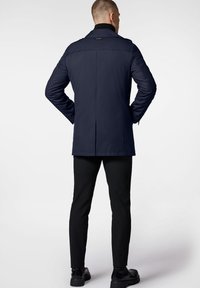 Manteau bleu marine au design structuré, doté d'un col montant, d'épaulettes et d'un dos droit, associé à un pantalon noir slim et des bottes.