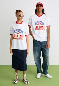 Witte T-shirts met rode rand tonen de tekst "ULTIMI ROMANTICI" in afbeelding. Een model draagt een denim rok en veterschoenen, terwijl het andere model jeans en sneakers draagt.