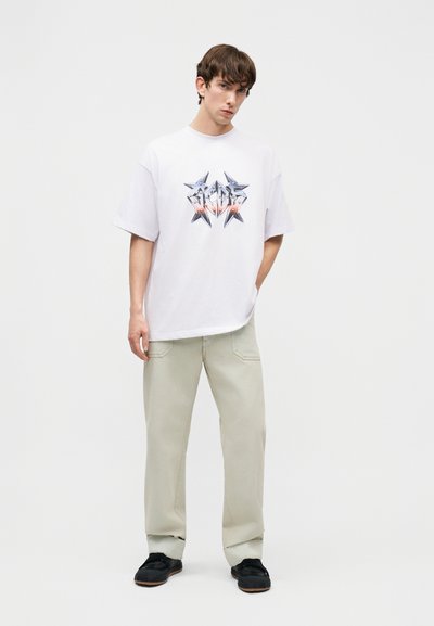 Νέος άνδρας στέκεται φορώντας λευκό φαρδύ μπλουζάκι με σχέδιο, ανοιχτό μπεζ παντελόνι και μαύρα αθλητικά παπούτσια, μπροστά σε απλό λευκό φόντο.