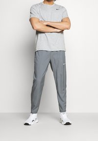 Mann in grauem Nike-T-Shirt und Hose mit weißen Nike-Sneakern, steht mit verschränkten Armen vor einem schlichten Hintergrund.