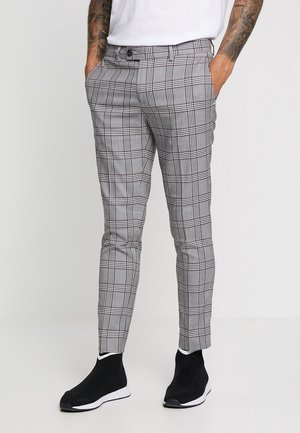 Mann trägt slim-fit graue karierte Hose, weißes Hemd und schwarze Sneaker im Sockendesign, steht mit den Händen in den Taschen.