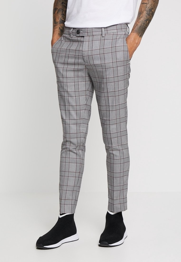 Homme portant un pantalon slim gris à carreaux, une chemise blanche et des baskets noires type chaussette, debout les mains dans les poches.