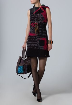 Mujer vestida con un vestido sin mangas de patrones rojo y negro, medias negras, tacones negros, llevando un gran bolso morado con un diseño azul.