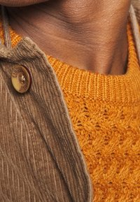 Gros plan sur un cou portant un pull orange en tricot texturé sous une veste en velours côtelé marron avec un bouton de marque.
