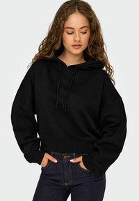 Felpa nera corta con cappuccio regolabile, maniche ampie e polsini a coste. Abbinata a jeans di denim blu scuro con chiusura a bottone.