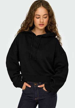 Felpa nera corta con cappuccio regolabile, maniche ampie e polsini a coste. Abbinata a jeans di denim blu scuro con chiusura a bottone.