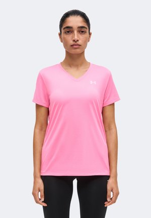 Sötét hajú nő konttyal, rózsaszín V-nyakú Under Armour pólót és fekete leggingset visel, előre néz homogén háttér előtt.